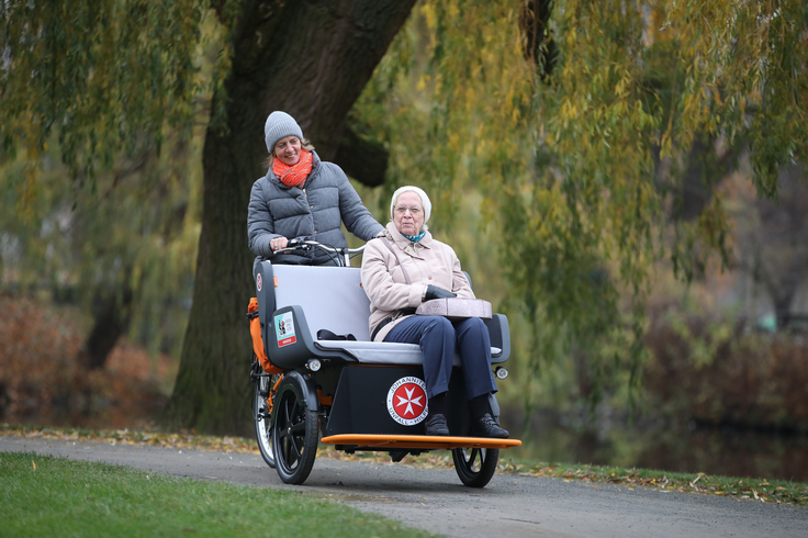 Eine Frau fährt eine ältere Dame in einem Park in einer Rikscha spazieren. Beide freuen sich sehr.