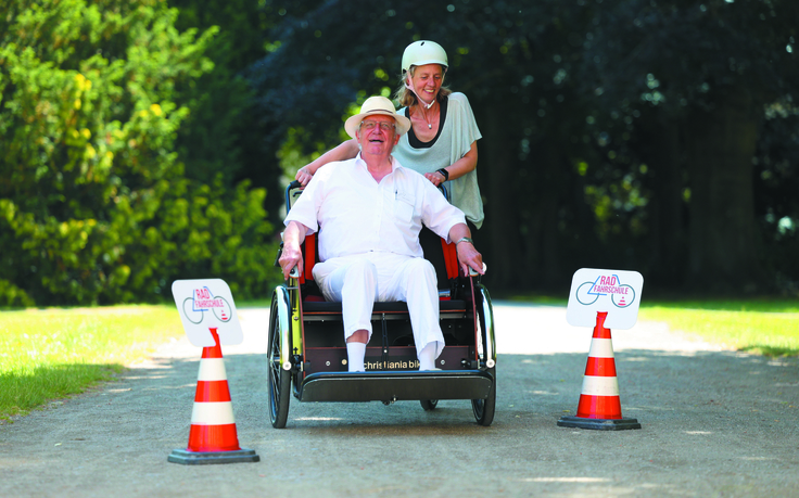 Ein älterer Herr wird in einer Rikscha von einer Frau durch einen Park gefahren. Sie fahren zwischen 2 Pilonen hindurch und haben Spaß!