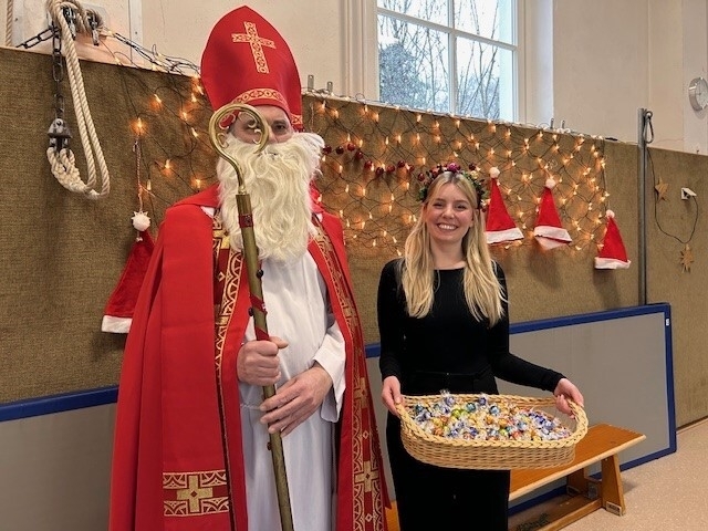 Der Nikolaus und Frau Musäus mit einem Korb voller Schokoladenkugeln vor einer geschmückten und beleuchteten Wand in der Gymnastikhalle im LBZB.