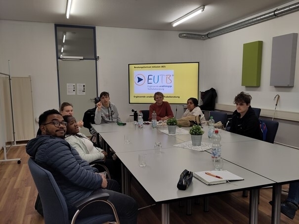 Eine Gruppe Schüler und Schülerinnen sitzen mit einer Teilhabe-Beraterin am Tisch in der Beratungsstelle “Mittendrin”. Im Hintergrund sieht man eine Power-Point-Präsentation über das Thema EUTB.