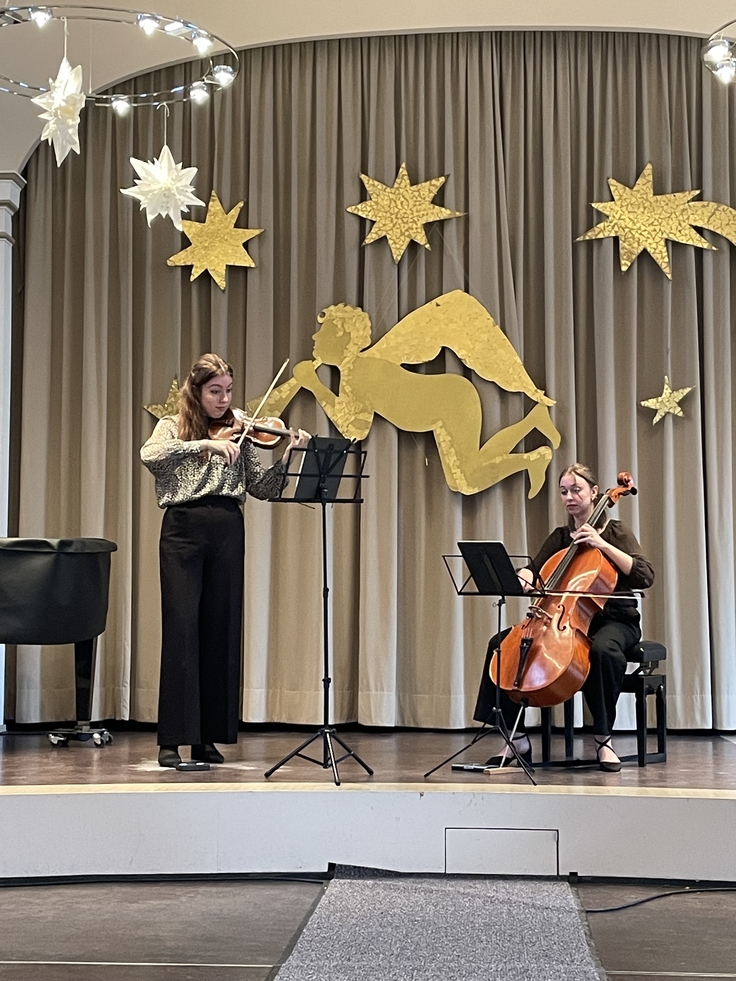 Eine Musikerin mit einer Geige und eine Musikerin mit einem Cello spielen auf der Bühne der weihnachtlich geschmückten Aula.