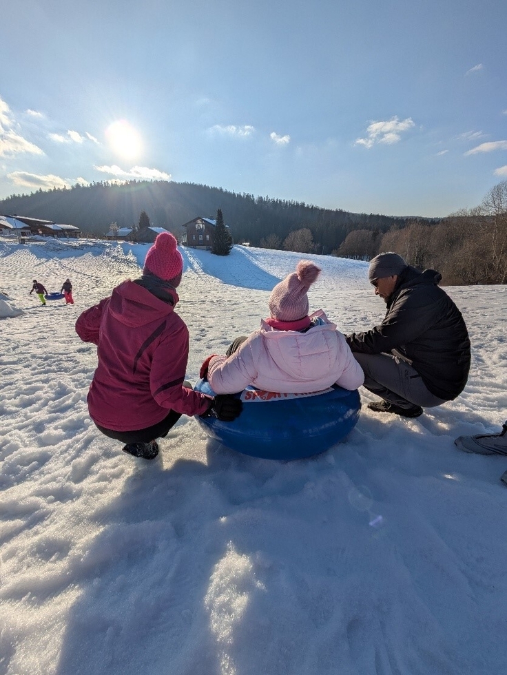 Frau Seelig und Herr Purevdorj hocken neben einer Schülerin, die bereits auf dem Rodelreifen sitzt und wartet, bis der Weg frei ist. Die drei Personen sind von hinten fotografiert. Unten am Berg sind noch zwei Schülerinnen zu sehen, die den zweiten Rei