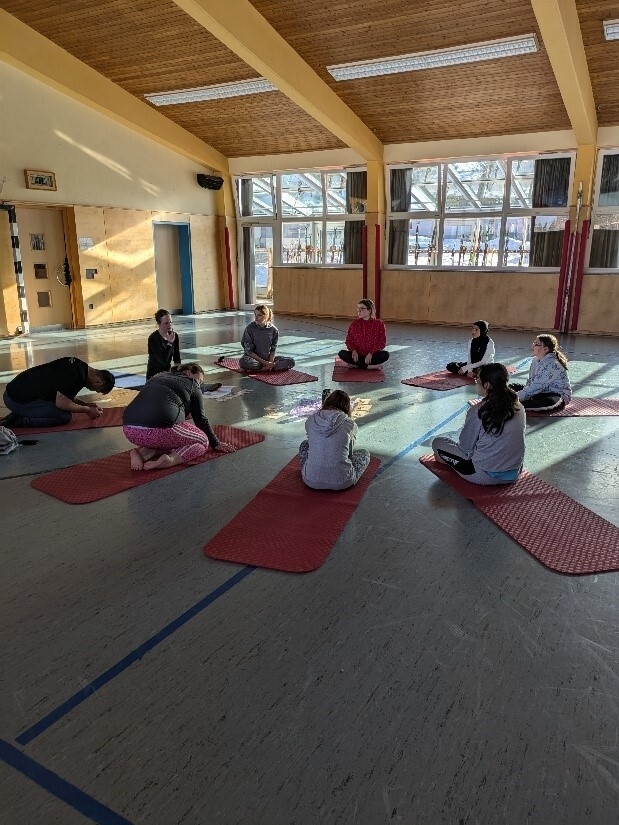 Die Schülerinnen und Lehrer*innen sitzen im Kreis auf Yogamatten in der Sporthalle und warten auf den Beginn der Yogaeinheit.