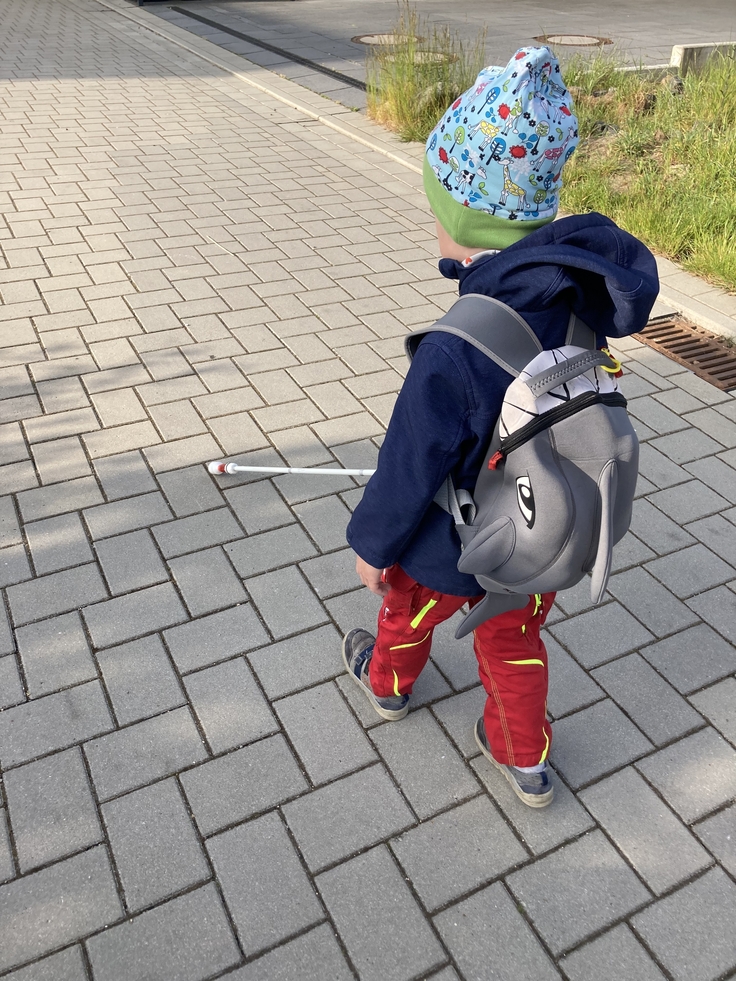 Auf dem Bild geht ein kleiner Junge mit einem Langstock den gepflasterten Weg entlang. Er trägt eine Mütze, einen Anorak und einen Rucksack. Er ist von hinten zu sehen.