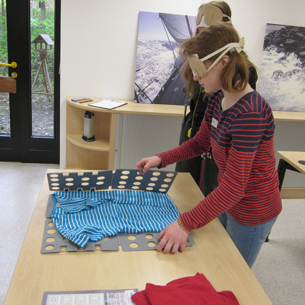 Svea legt ein T-shirt mit Unterstützung einer Falthilfe zusammen.