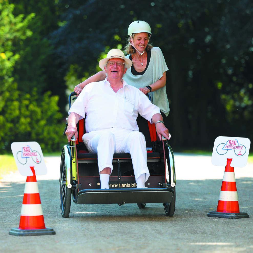 Ein älterer Herr wird in einer Rikscha von einer Frau durch einen Park gefahren. Sie fahren zwischen 2 Pilonen hindurch und haben Spaß!