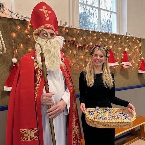 Der Nikolaus und Frau Musäus mit einem Korb voller Schokoladenkugeln vor einer geschmückten und beleuchteten Wand in der Gymnastikhalle im LBZB.