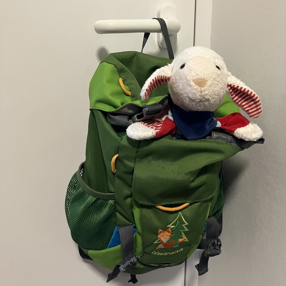 An einer weißen Türklinke hängt ein dunkelgrüner Rucksack. Aus der oberen Öffnung schauen der Kopf und die Vorderbeine eines Kuscheltier-Schafs heraus.